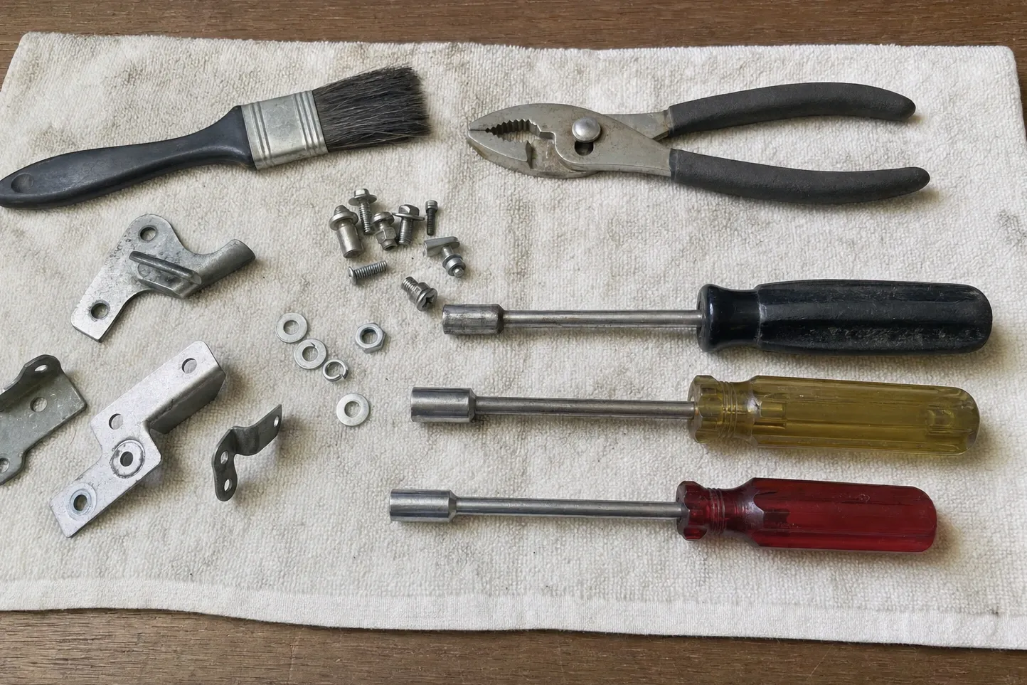 A soft dusting brush and slip-joint pliers beside three hinge plates, a small formed clip, washers, nuts and three nut drivers